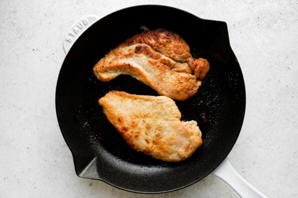 How to make Meyer Lemon Chicken Piccata, step 4: Brown the chicken. Two browned chicken cutlets sit in a white Staub cast iron skillet. The skillet sits atop a creamy white textured surface.