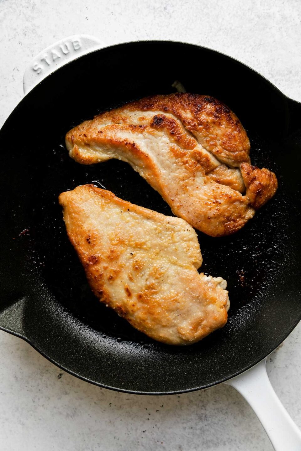 How to make Meyer Lemon Chicken Piccata, step 4: Brown the chicken. Two browned chicken cutlets sit in a white Staub cast iron skillet. The skillet sits atop a creamy white textured surface.