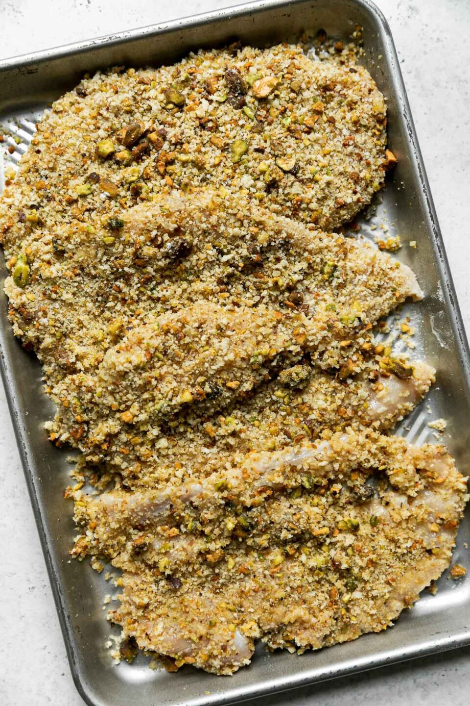 An aluminum baking sheet is filled with Parmesan Pistachio Crusted Chicken cutlets that have been breaded and prepared for cooking. The baking sheet sits atop a creamy white textured surface.