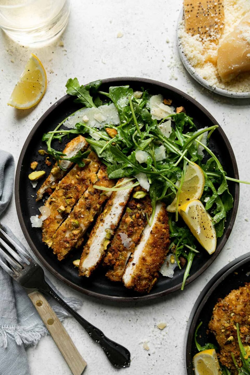 A serving of parmesan pistachio crusted chicken has been sliced and plated on a black ceramic dinner plate alongside a mixed greens salad dressed with more pistachios and parmesan and a light oil dressing. Two lemon wedges sit atop the salad. A silver fork and knife with a wooden handle, a clear glass filled with white wine, a small ceramic plate filled with whole blocks of Parmigiano Reggiano and grated Parmigiano Reggiano, a single lemon wedge, a light blue linen napkin, and another plate of pistachio chicken surround the plate at center. All items sit atop a creamy white textured surface.