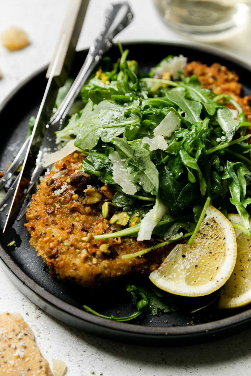 A side angle shot of Parmigiano Reggiano pistachio crusted chicken on a large black dinner plate with a green salad, two lemon wedges, and a silver fork and knife with a wooden handle. The plate has been garnished with black pepper and shaved parmesan. A clear glass filled with white wine and a block of parmesan cheese surround the plate. All items sit atop a creamy white textured surface.