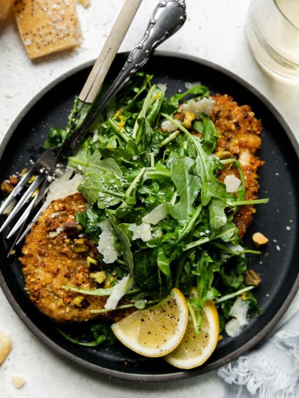 Parmigiano Reggiano pistachio crusted chicken on a large black dinner plate with a green salad, two lemon wedges, and a silver fork and knife with a wooden handle. The plate has been garnished with black pepper and shaved parmesan. A light blue linen napkin, a clear glass filled with white wine, and a Parmigiano Reggiano rind surround the plate. All items sit atop a creamy white textured surface.