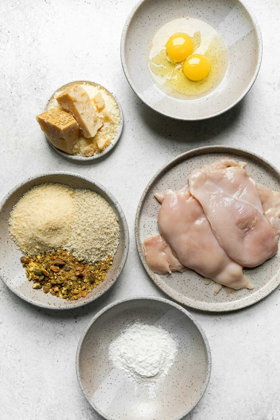 Parmesan pistachio crusted chicken ingredients are arranged in speckled gray ceramic bowls and plates a top a creamy white texutred surface: boneless, skinless chicken breasts, all-purpose flour, eggs, pistachios, panko breadcrumbs, and Parmigiano Reggiano.