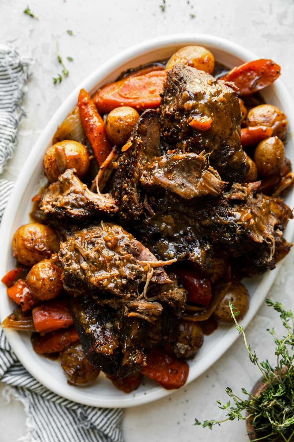 Braised pot roast with vegetables are plated for serving atop a white platter. The platter sits atop a creamy white textured surface with a blue and white striped linen napkin and a small wooden pinch bowl filled with fresh thyme surrounding the platter.