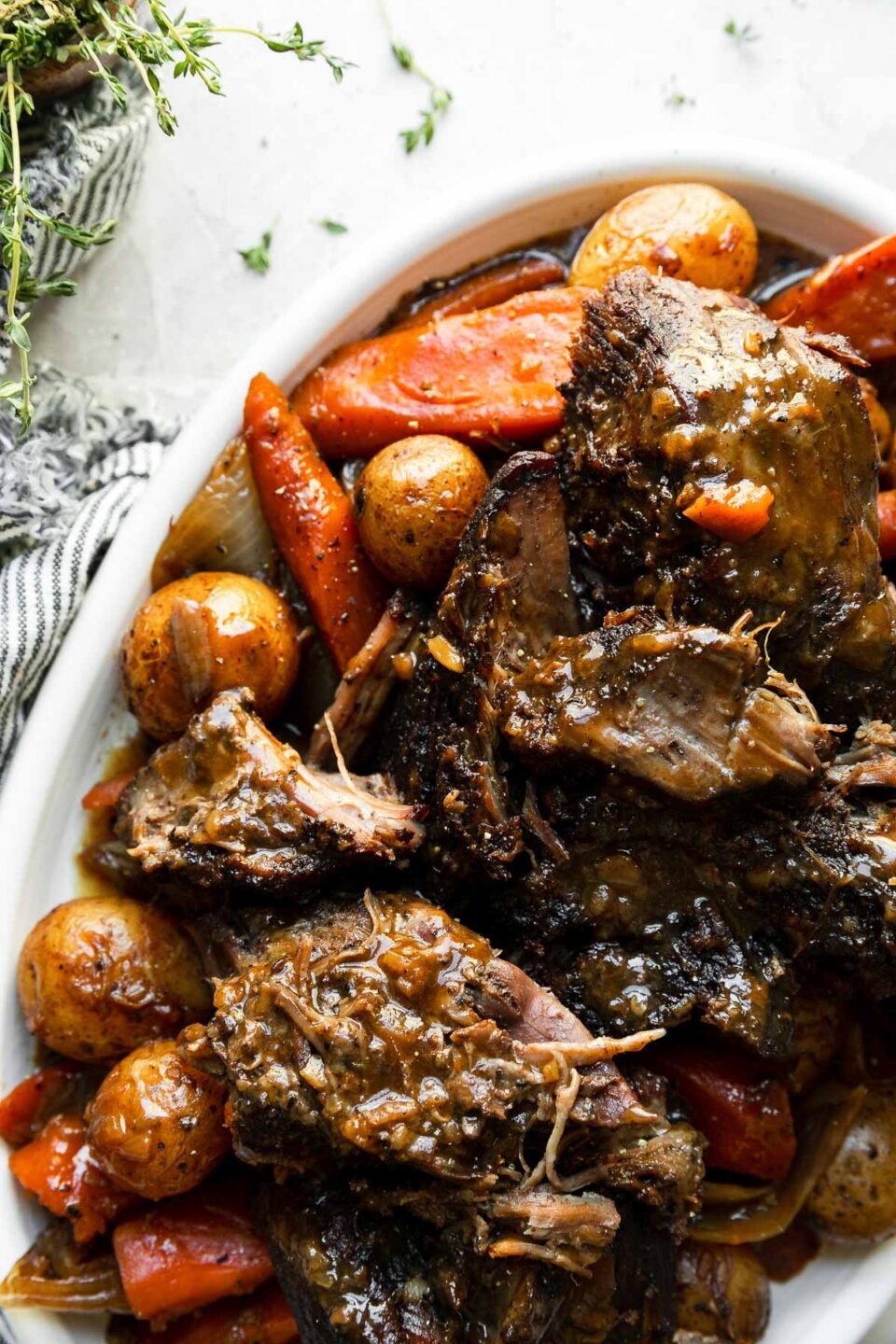 A close up of braised pot roast with vegetables are plated for serving atop a white platter. The platter sits atop a creamy white textured surface with a blue and white striped linen napkin and a small wooden pinch bowl filled with fresh thyme surrounding the platter.