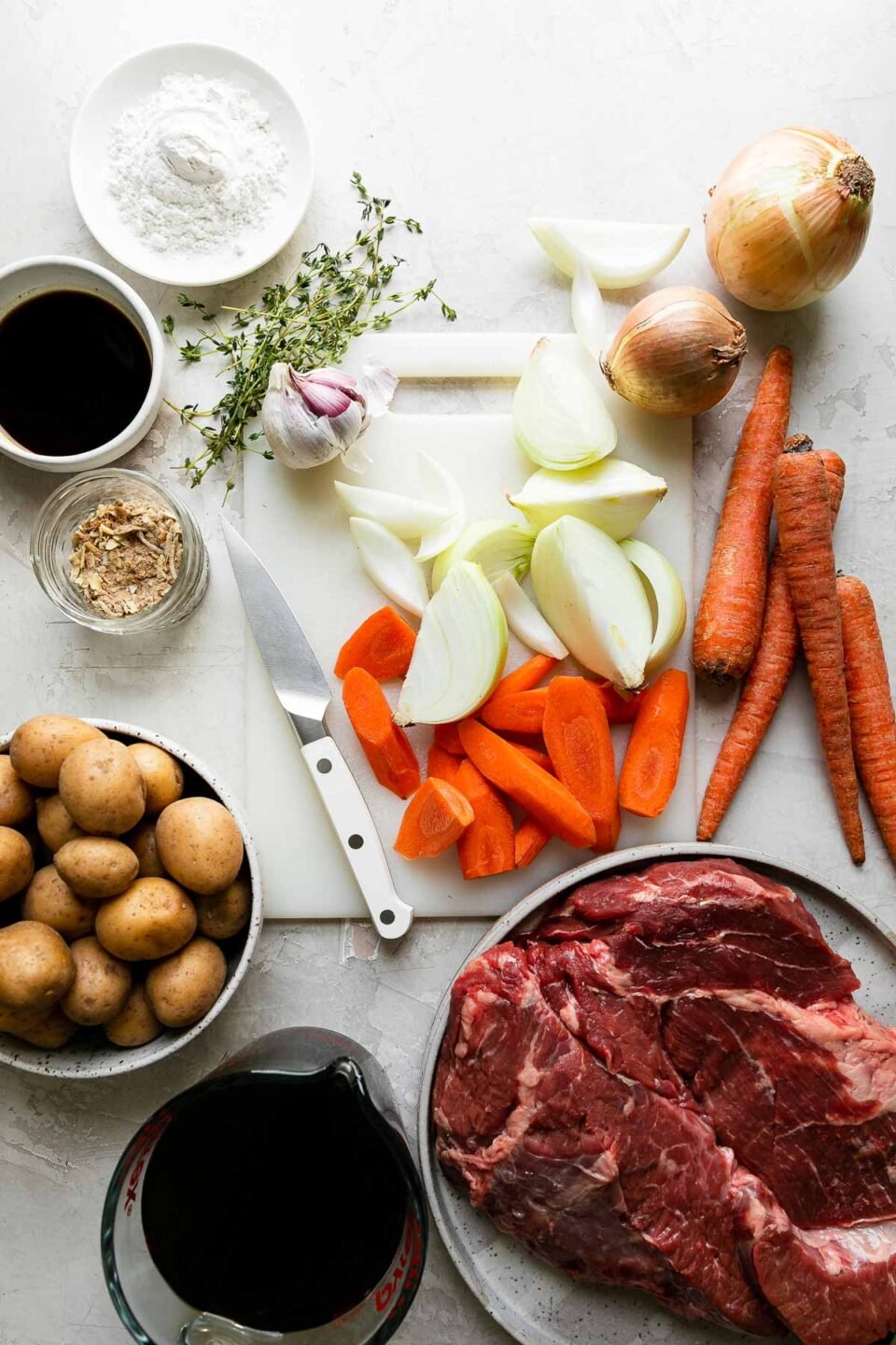 Mom's Extra-Veggies Pot Roast with vegetables ingredients arranged on na creamy white textured surface: boneless beef chuck roast, all-purpose flour, olive oil, baby gold potatoes, carrots, yellow onions, garlic, beef stock, Worcestershire sauce, and dry onion soup mix.