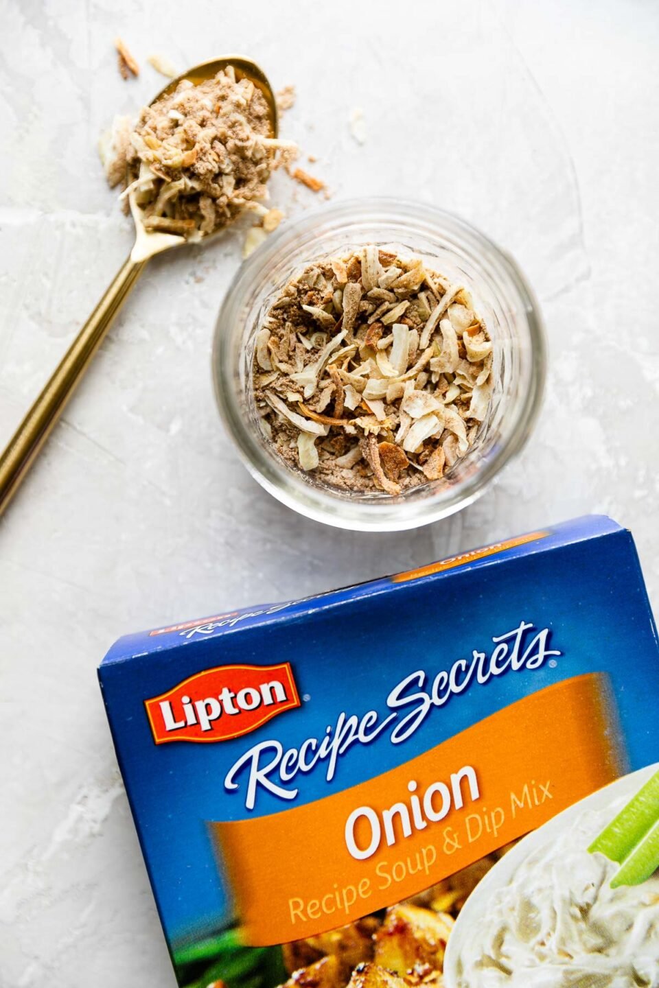 A package of Lipton dry Onion Soup Mix lays flat on a creamy white textured surface. Above the box rests a small mason jar filled with onion soup mix and a spoonful of the mix fills a small gold spoon that rests alongside the mason jar. This is one of the secret ingredients for making this dutch oven pot roast recipe.
