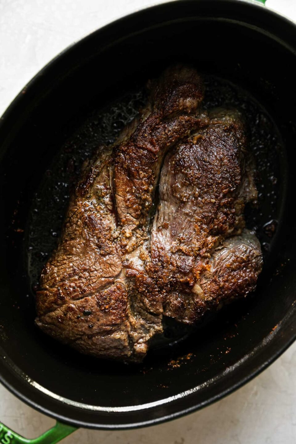 How to make braised pot roast, Step 2: Brown the chuck roast. Browned chuck roasts rests at the bottom of an oval green Staub dutch oven. The dutch oven sits atop a creamy white textured surface.