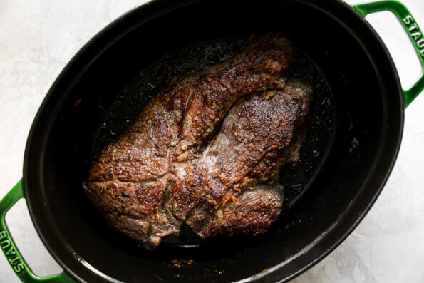 How to make braised pot roast, Step 2: Brown the chuck roast. Browned chuck roasts rests at the bottom of an oval green Staub dutch oven. The dutch oven sits atop a creamy white textured surface.