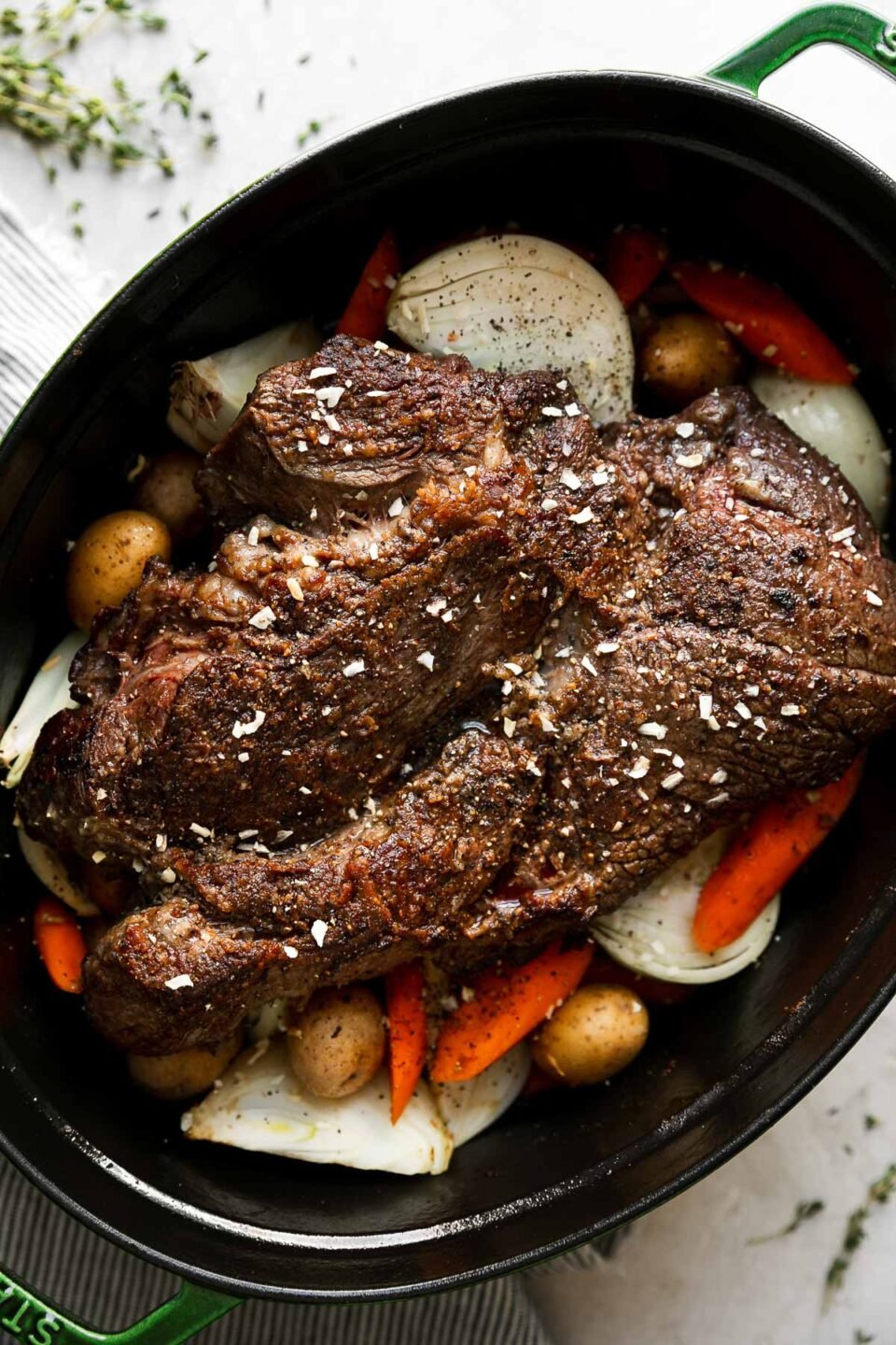 How to make pot roast with vegetables, step 3: Assemble the braised pot roast. An oval green Staub cast iron dutch oven is filled with a browned chuck roast. Halved or roughly chopped baby potatoes, carrots, and yellow onion have been added to the dutch oven surrounding the roast and beef stock, Worcestershire sauce, and dry onion soup mix have also been added over top. The dutch oven sits atop a creamy white textured surface with fresh sprigs of thyme and a blue and white striped linen napkin surrounding the dutch oven.