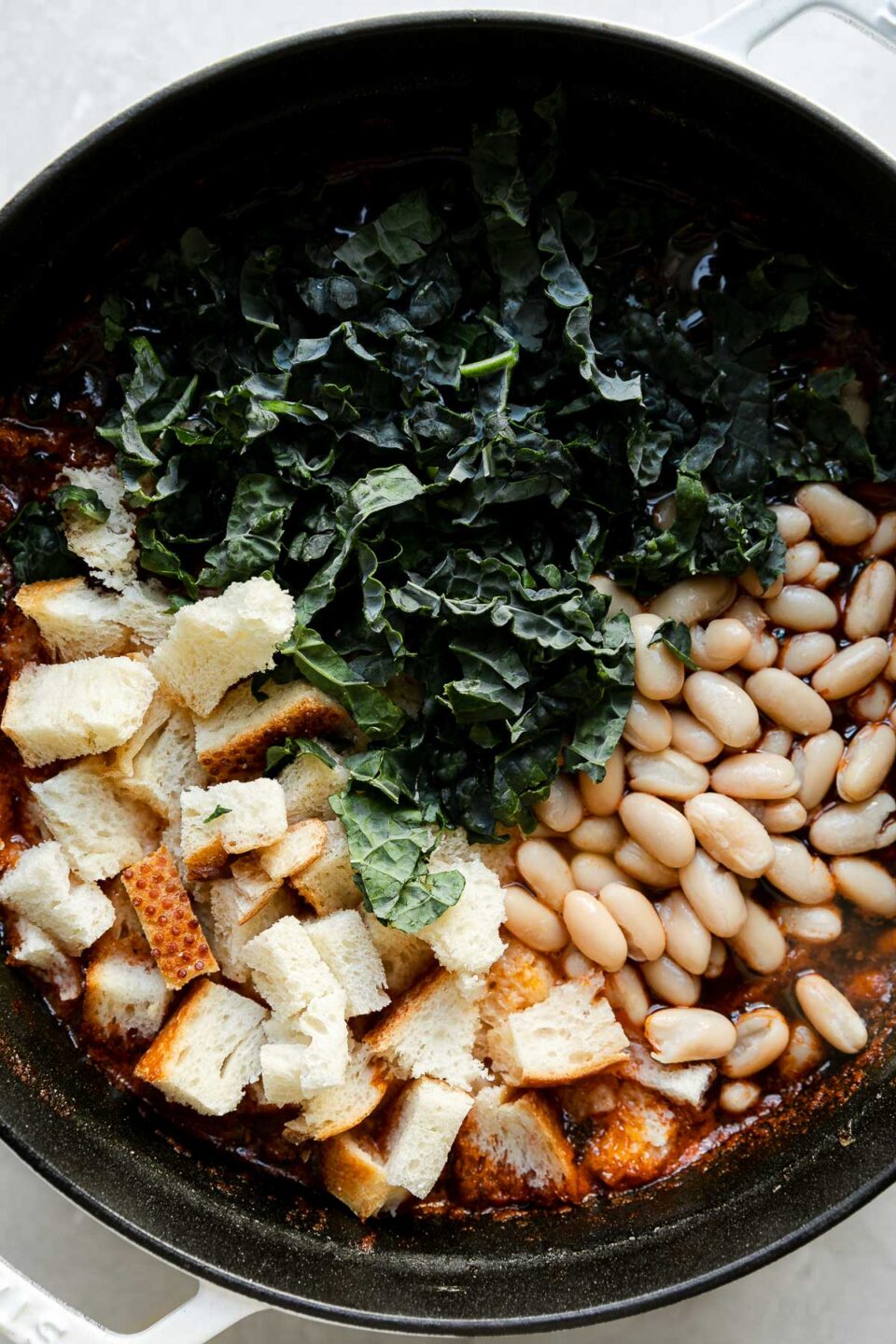 A close up of shredded kale, white cannellini beans, and torn bread are added to Ribollita soup broth within a white Staub cocotte. The cocotte sits atop a creamy white textured surface.