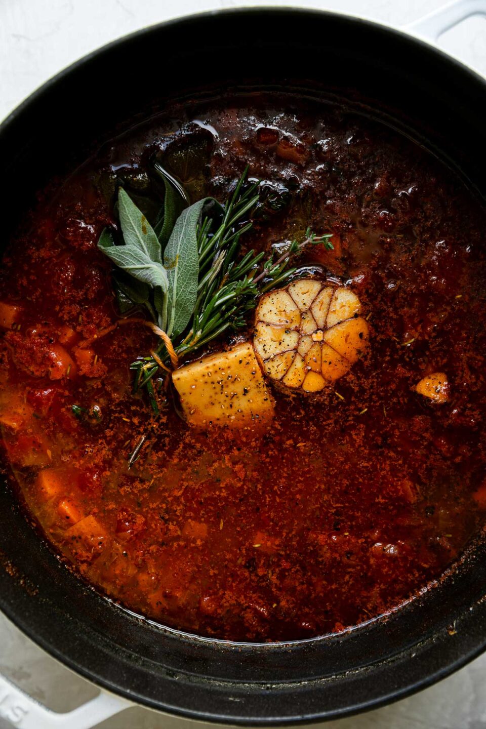 Ribollita soup broth fills a white Stab cocotte. Garlic, bay leaves, parmesan rind, & fresh herbs have been added to the broth to flavor. The cocotte sits atop a creamy white textured surface.