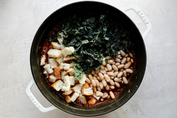 Shredded kale, white cannellini beans, and torn bread are added to Ribollita soup broth within a white Staub cocotte. The cocotte sits atop a creamy white textured surface.