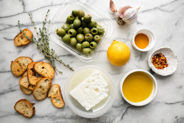 Heavenly Baked Feta with Olives ingredients arranged a white and gray marble surface: feta cheese, olive oil, honey, garlic, fresh thyme, crushed red pepper flakes, Castelvetrano olives, lemon, and crostini for serving.