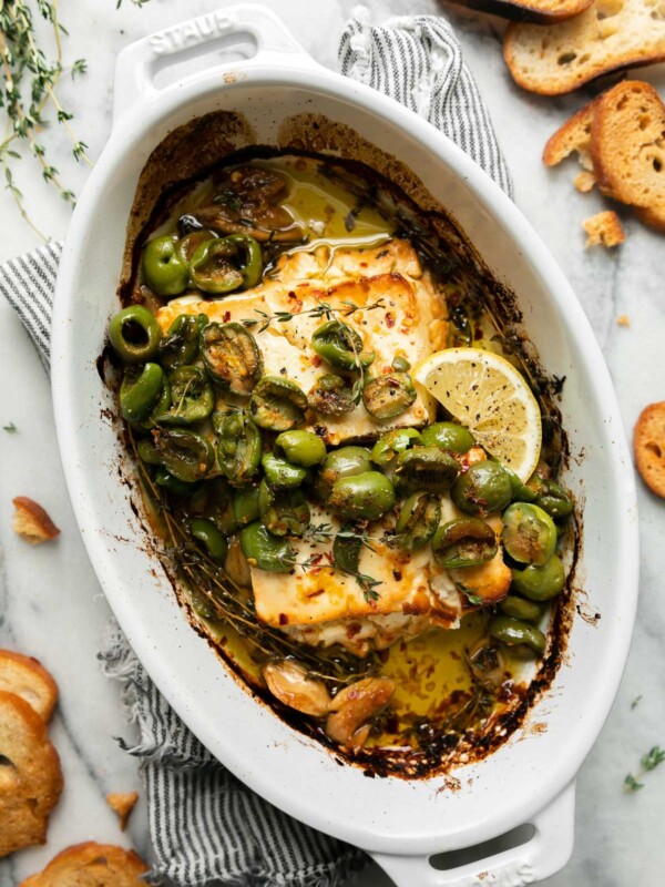 Finished baked feta with Castelvetrano olives is topped with fresh sprigs of thyme and a lemon wedge in a small white Staub baking dish. The baking dish sits atop a blue and white striped linen napkin that sits atop a gray and white marble surface. Pieces of crostini and sprigs of fresh thyme surround the baking dish.