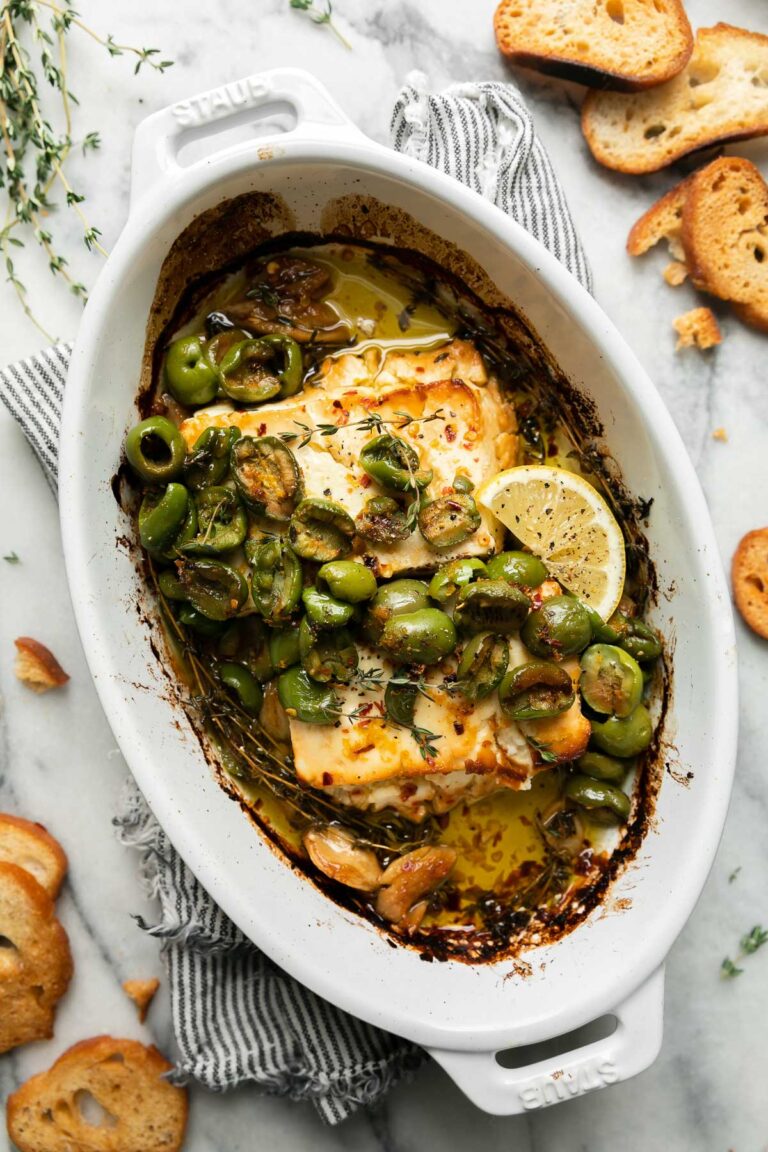 Finished baked feta with Castelvetrano olives is topped with fresh sprigs of thyme and a lemon wedge in a small white Staub baking dish. The baking dish sits atop a blue and white striped linen napkin that sits atop a gray and white marble surface. Pieces of crostini and sprigs of fresh thyme surround the baking dish.