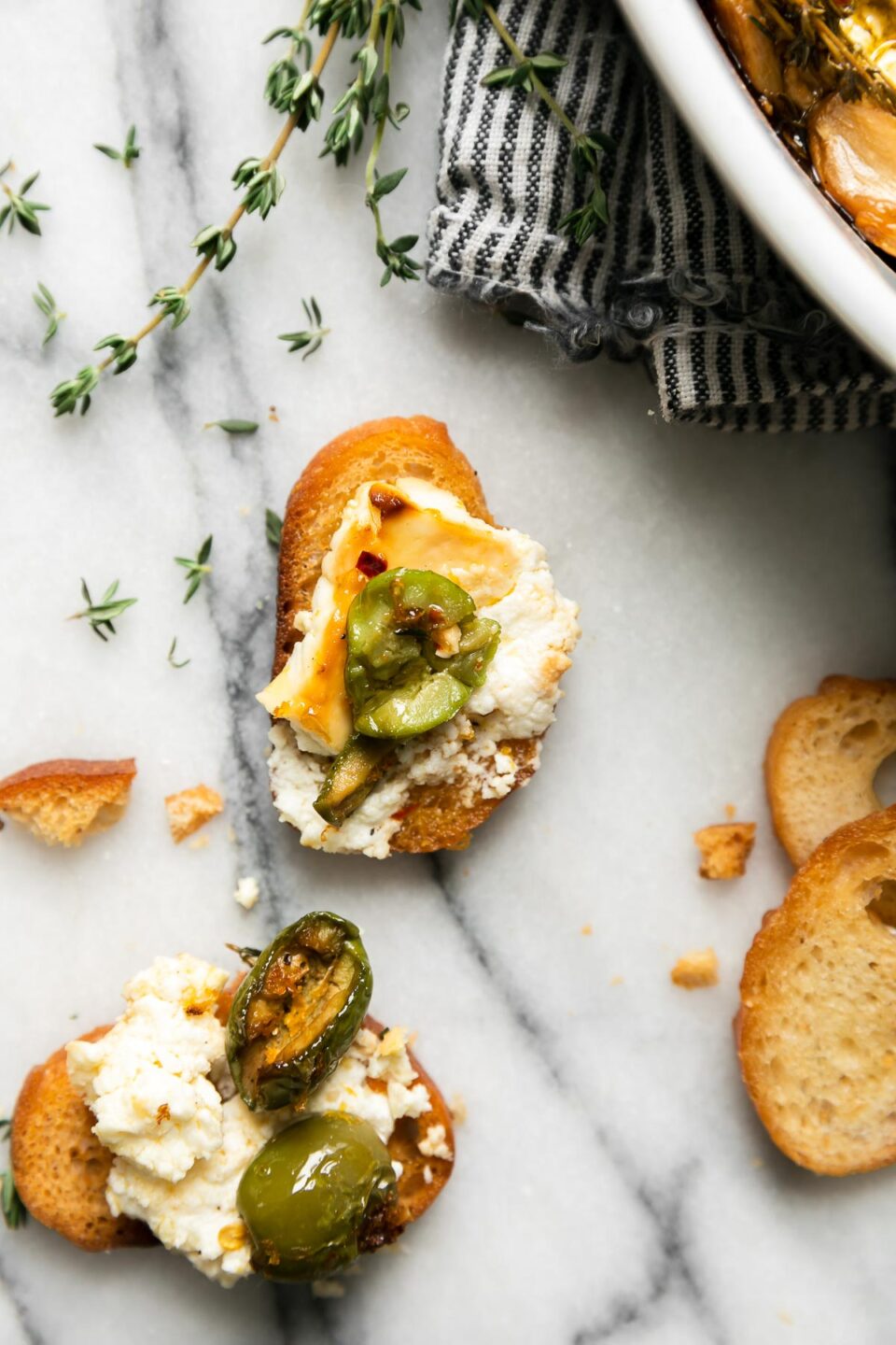 A side angle and close up shot of two crostinis topped with baked feta with Castelvetrano olives. A small white Staub baking dish is filled with baked feta with olives that rests behind the crostini atop a blue and white striped linen napkin that sits atop a gray and white marble surface. Sprigs of fresh thyme and crostini pieces surround the baking dish and prepared crostini.
