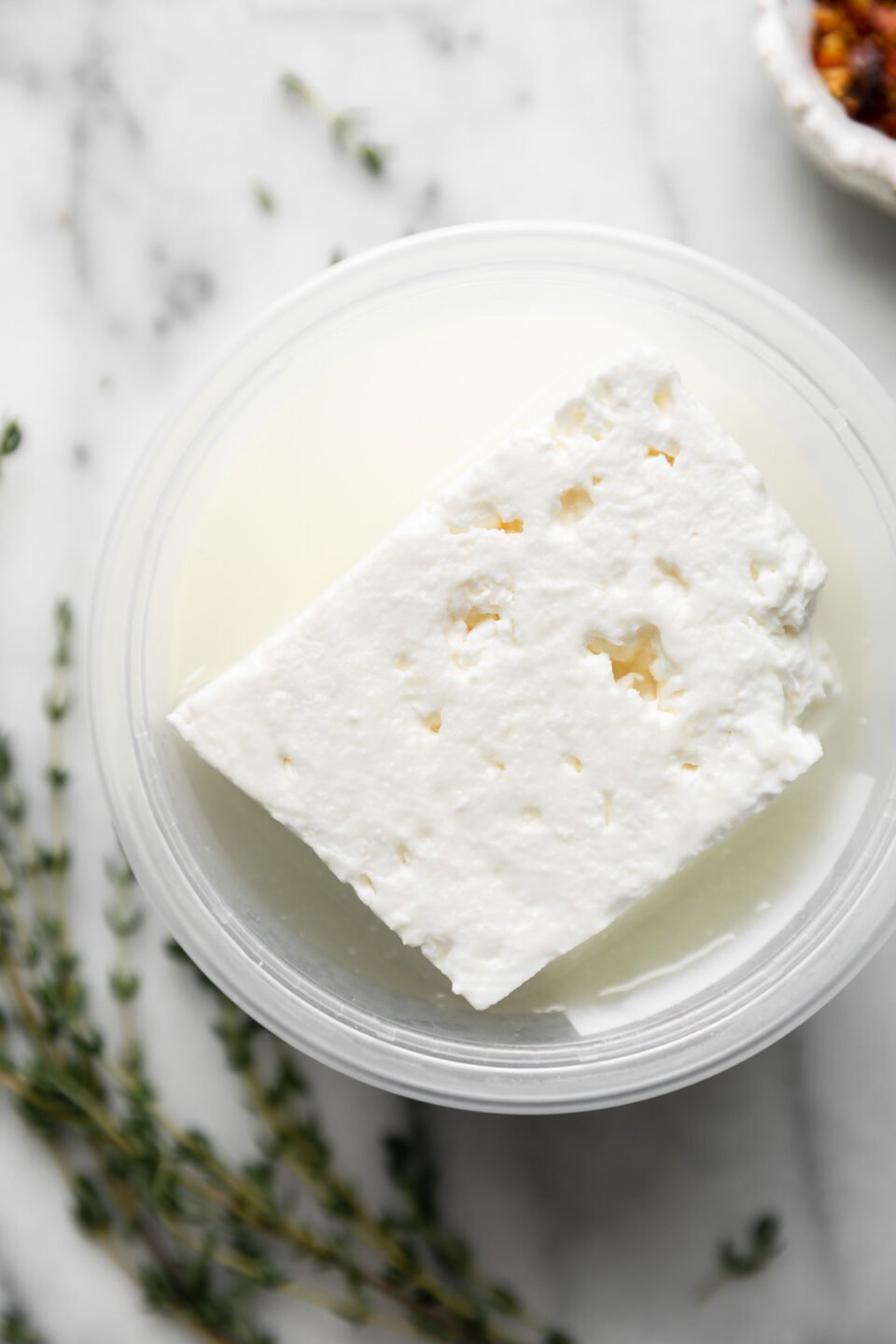 A block of feta cheese in brine rests inside a plastic deli container. The container sits atop a gray and white marble surface. A small pinch bowl filled with crushed red pepper flakes and fresh sprigs of thyme rest alongside the feta.