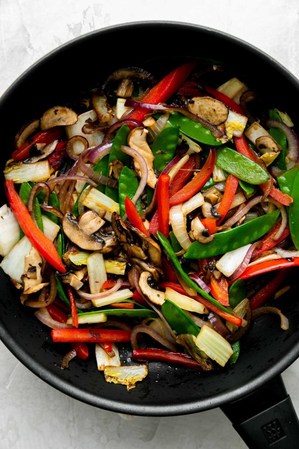 How to make beef and vegetable stir fry, step 3: Stir fry the vegetables. Another batch of stir fry veggies including red onion, red bell pepper, mushrooms, napa cabbage, and snow peas cook in oil in a Zwilling Madura Nonstick Skillet. The skillet sits atop a creamy white textured surface.