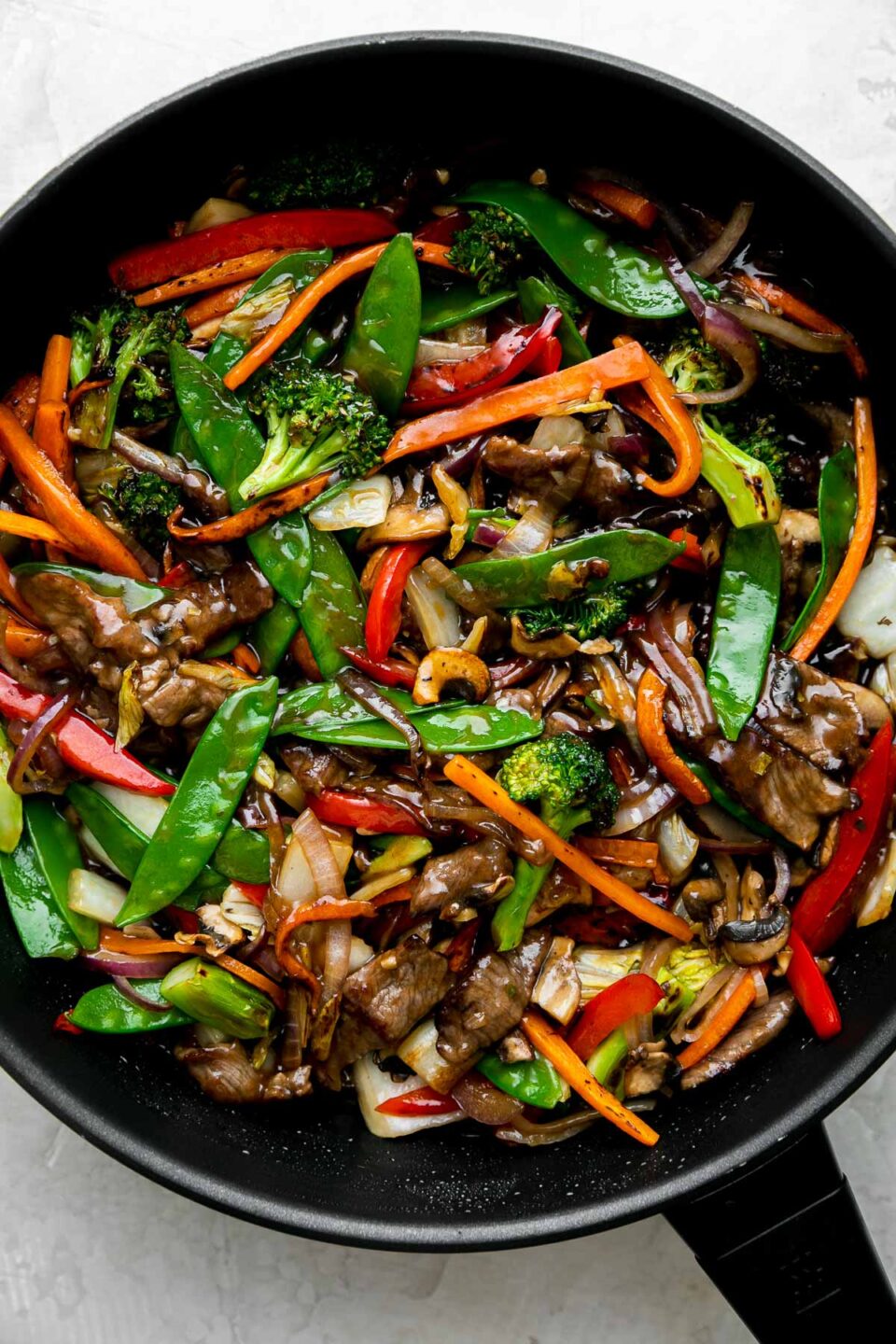 How to make beef and vegetable stir fry, step 4: Sear the beef & finish the stir fry. Finished beef vegetable stir fry fills a black Zwilling Madura Nonstick Skillet. The beef and vegetables are glossy with a coating of homemade stir fry sauce. The skillet sits atop a creamy white textured surface.