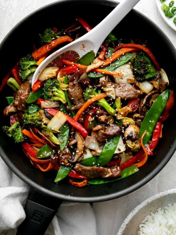Finished beef vegetable stir fry fills a black Zwilling Madura Nonstick Skillet. The beef and vegetables are glossy with a coating of homemade stir fry sauce. The skillet sits atop a creamy white textured surface with a light gray linen napkin tucked underneath. A white Chinese soup spoon rests inside of the skillet for serving, while a small white plate filled with sliced green onion and small gray speckled ceramic bowl filled with rice rest alongside. The stir fry is garnished with toasted sesame seeds.
