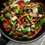Finished beef vegetable stir fry fills a black Zwilling Madura Nonstick Skillet. The beef and vegetables are glossy with a coating of homemade stir fry sauce. The skillet sits atop a creamy white textured surface with a light gray linen napkin tucked underneath. A white Chinese soup spoon rests inside of the skillet for serving, while a small white plate filled with sliced green onion and small gray speckled ceramic bowl filled with rice rest alongside. The stir fry is garnished with toasted sesame seeds.