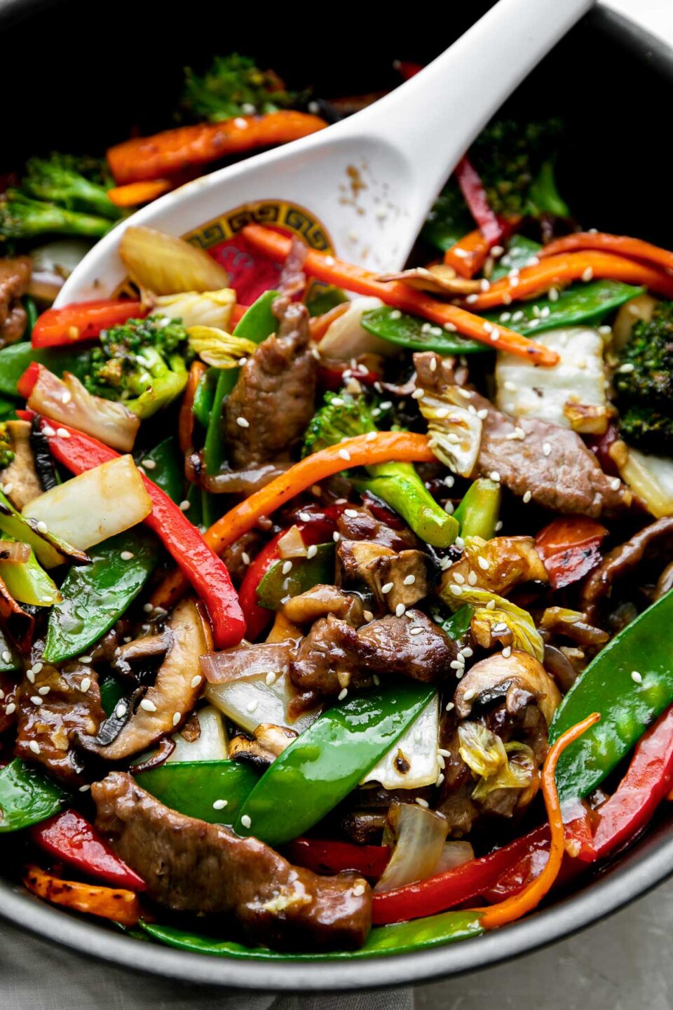 A close up shot of finished beef vegetable stir fry fills a black Zwilling Madura Nonstick Skillet. The beef and vegetables are glossy with a coating of homemade stir fry sauce. The skillet sits atop a creamy white textured surface with a light gray linen napkin tucked underneath. A white Chinese soup spoon rests inside of the skillet for serving.