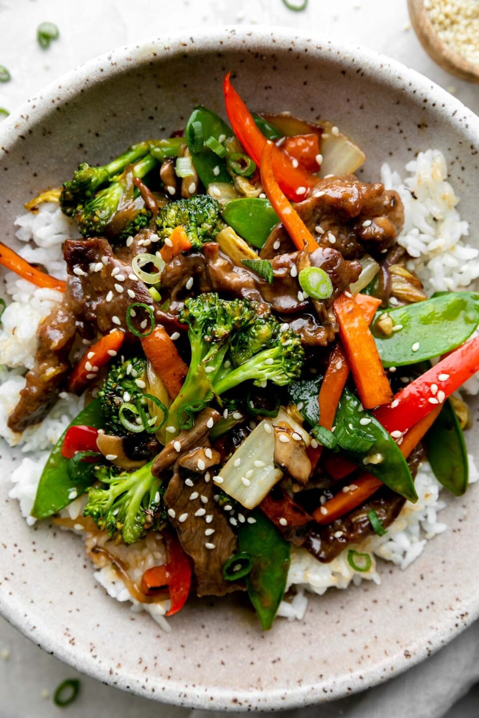 A serving of beef and vegetable stir fry fills a gray speckled ceramic bowl atop a bed of white rice. The beef and veggie stir fry has been garnished with toasted sesame seeds and sliced green onion. The bowl sits atop a creamy white textured surface with a light gray linen napkin is tucked alongside the bowl. A small wooden pinch bowl filled with toasted sesame seeds and loosely sprinkled sliced green onion surround the bowl of stir fry.