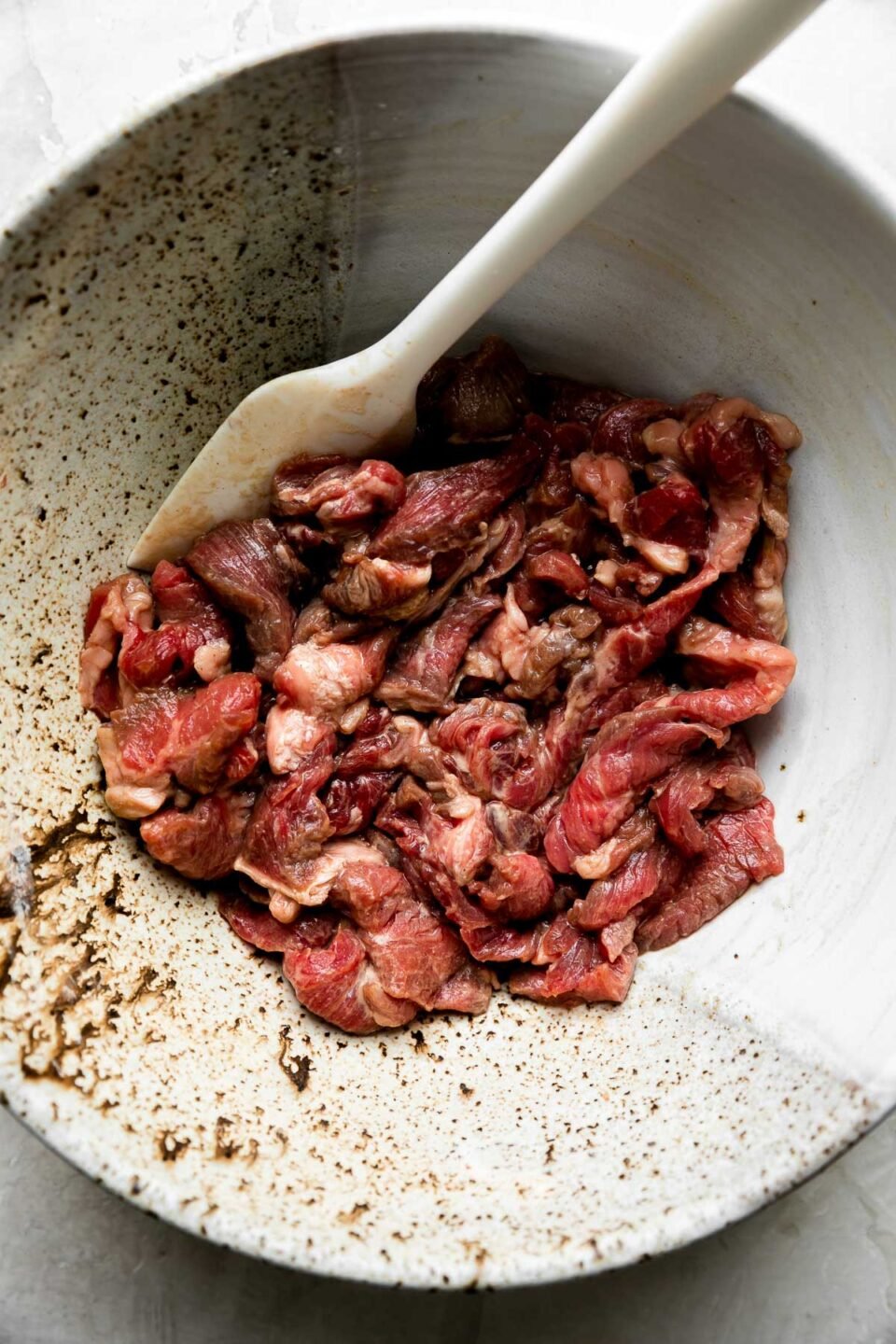 Thinly sliced top sirloin steak marinates in a gray speckled ceramic bowl filled with cornstarch, soy sauce, and rice wine for a beef and veggie stir fry. A white rubber spatula rests inside of the bowl for mixing and the bowl sits atop a creamy white textured surface.