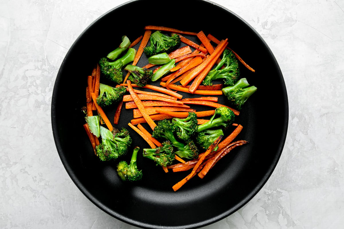 Broccoli cut into bite-sized florets and carrot sliced into strips cook in a black Zwilling Madura Nonstick Skillet in oil. The skillet sits atop a creamy white textured surface.