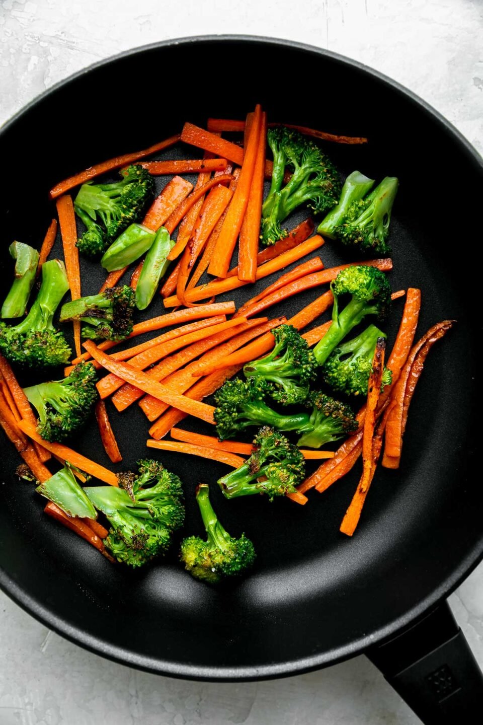 Broccoli cut into bite-sized florets and carrot sliced into strips cook in a black Zwilling Madura Nonstick Skillet in oil. The skillet sits atop a creamy white textured surface.