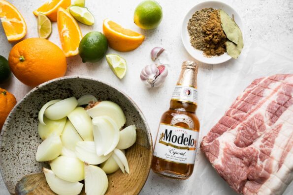 Beer braised carnitas ingredients arranged on a creamy white textured surface: boneless pork shoulder, garlic, yellow onions, navel oranges, limes, ground cumin, dried oregano, bay leaves, light beer of choice.