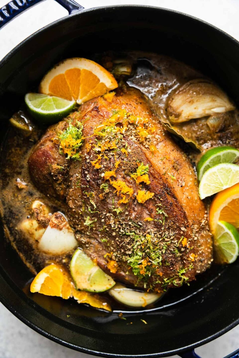 An overhead shot of how to make Dutch oven carnitas, step 3: Assemble the braise. A browned boneless pork shoulder sits inside of a dark navy Staub dutch oven for Dutch oven carnitas. The pork shoulder is surrounded by onions & fresh citrus and rests inside of a braising liquid of light beer. Orange zest, lime zest, ground cumin, dried oregano, & bay leaf have been sprinkled over top. The pot sits atop a creamy white textured surface.