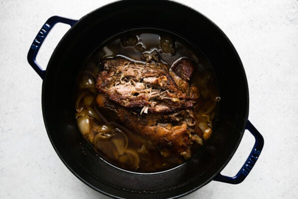 Cooked pork carnitas in a beer braise fill a dark navy Staub dutch oven. The braised carnitas have been slightly shredded on the top to check for doneness. The dutch oven sits atop a creamy white textured surface.
