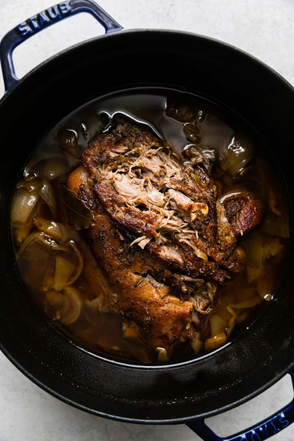 Cooked pork carnitas in a beer braise fill a dark navy Staub dutch oven. The braised carnitas have been slightly shredded on the top to check for doneness. The dutch oven sits atop a creamy white textured surface.