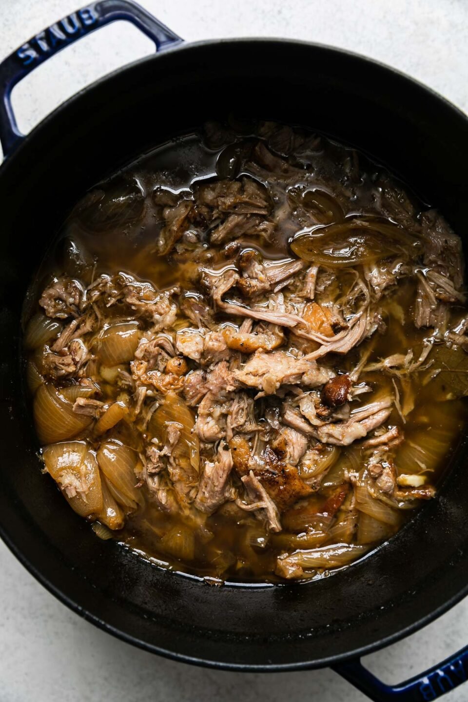 Cooked & shredded pork carnitas in a beer braise fill a dark navy Staub dutch oven. The dutch oven sits atop a creamy white textured surface.