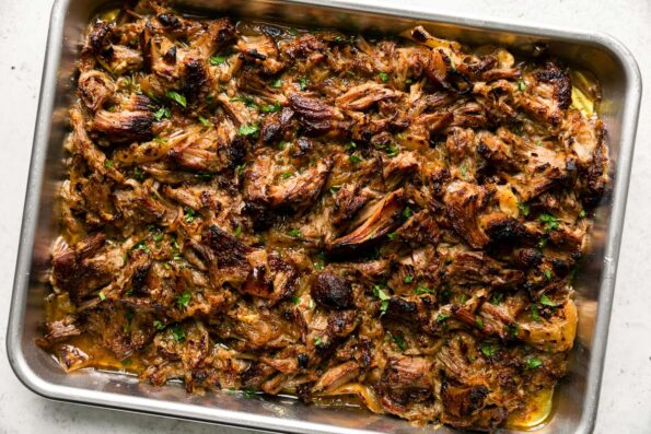 Cooked & shredded pork carnitas arranged on an aluminum baking sheet & broiled until golden brown with crisped edges. The baking sheet sits atop a creamy white textured surface and the beer braised carnitas have been garnished with fresh cilantro.