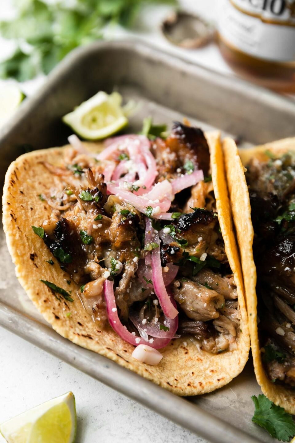 A side angle shot of beer braised carnitas tacos topped with pickled red onions, fresh chopped cilantro, queso fresco, with lime wedges on the side. The tacos sit atop a parchment paper lined aluminum baking sheet that sits atop a creamy white surface surrounded by lime wedges, a bouquet of fresh cilantro, a bottle of Modelo beer, and a loose beer bottle cap.