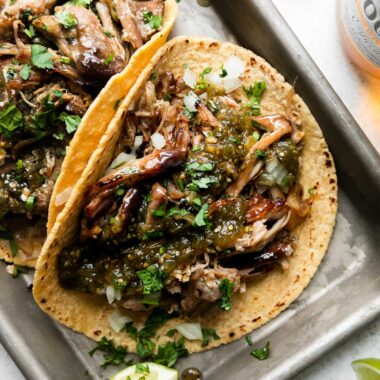 An overhead shot of beer braised carnitas tacos topped with salsa verde, fresh chopped cilantro, diced yellow onion, with lime wedges on the side. The tacos sit atop an aluminum baking sheet that sits atop a creamy white surface surrounded by lime wedges, a bottle of Modelo beer, and a loose beer bottle cap.
