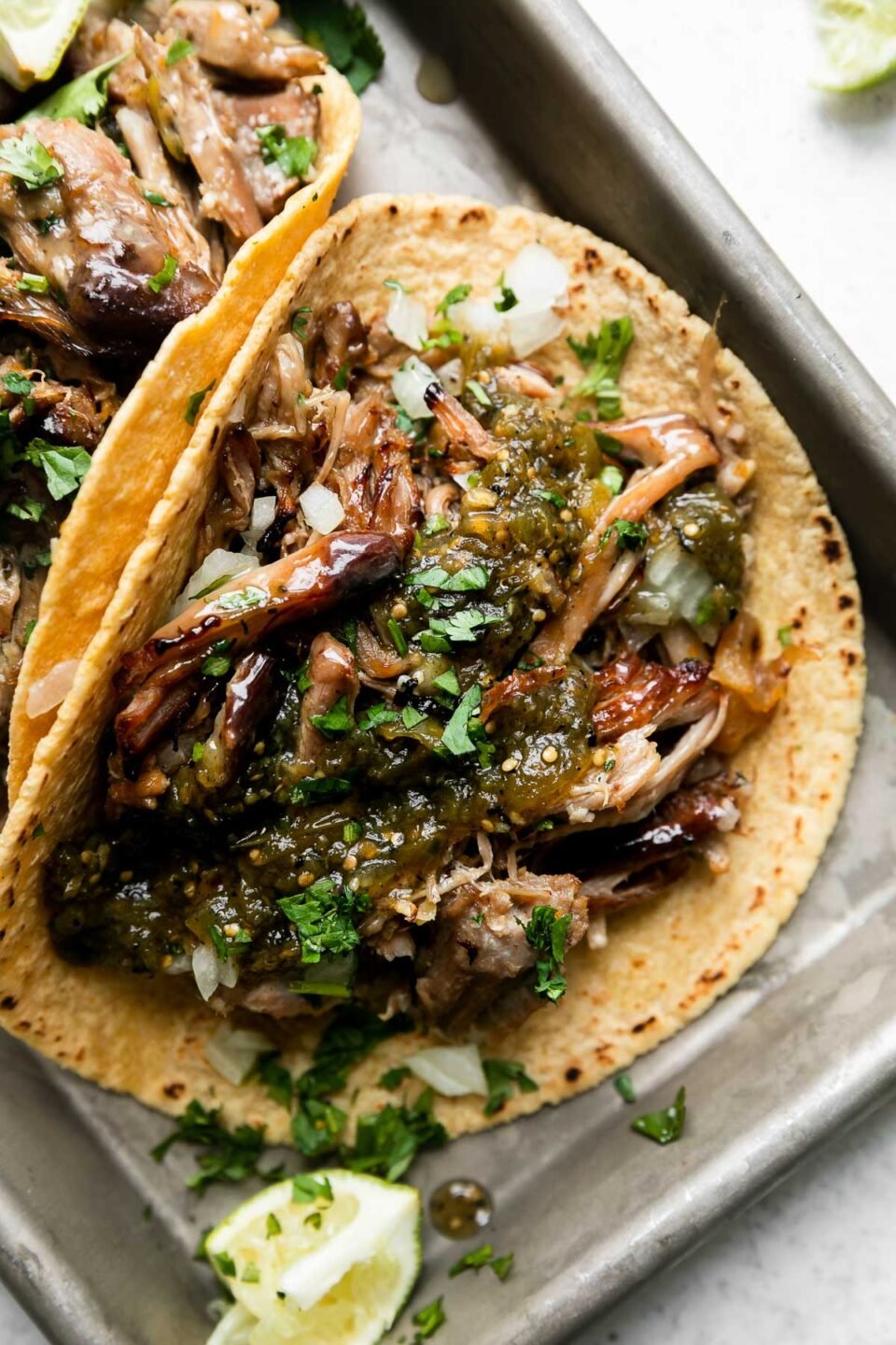 An overhead shot of beer braised carnitas tacos topped with salsa verde, fresh chopped cilantro, diced yellow onion, with lime wedges on the side. The tacos sit atop an aluminum baking sheet that sits atop a creamy white surface surrounded by loose lime wedges.
