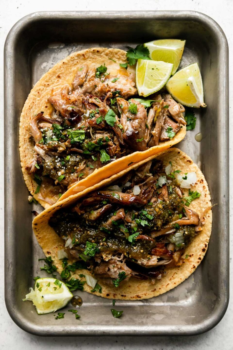 An overhead shot of beer braised carnitas tacos topped with salsa verde, fresh chopped cilantro, diced yellow onion, with lime wedges on the side. The tacos sit atop an aluminum baking sheet that sits atop a creamy white surface.