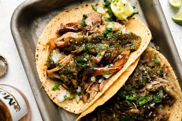 An overhead shot of beer braised carnitas tacos topped with salsa verde, fresh chopped cilantro, diced yellow onion, with lime wedges on the side. The tacos sit atop an aluminum baking sheet that sits atop a creamy white surface surrounded by lime wedges, a bottle of Modelo beer, and a loose beer bottle cap.