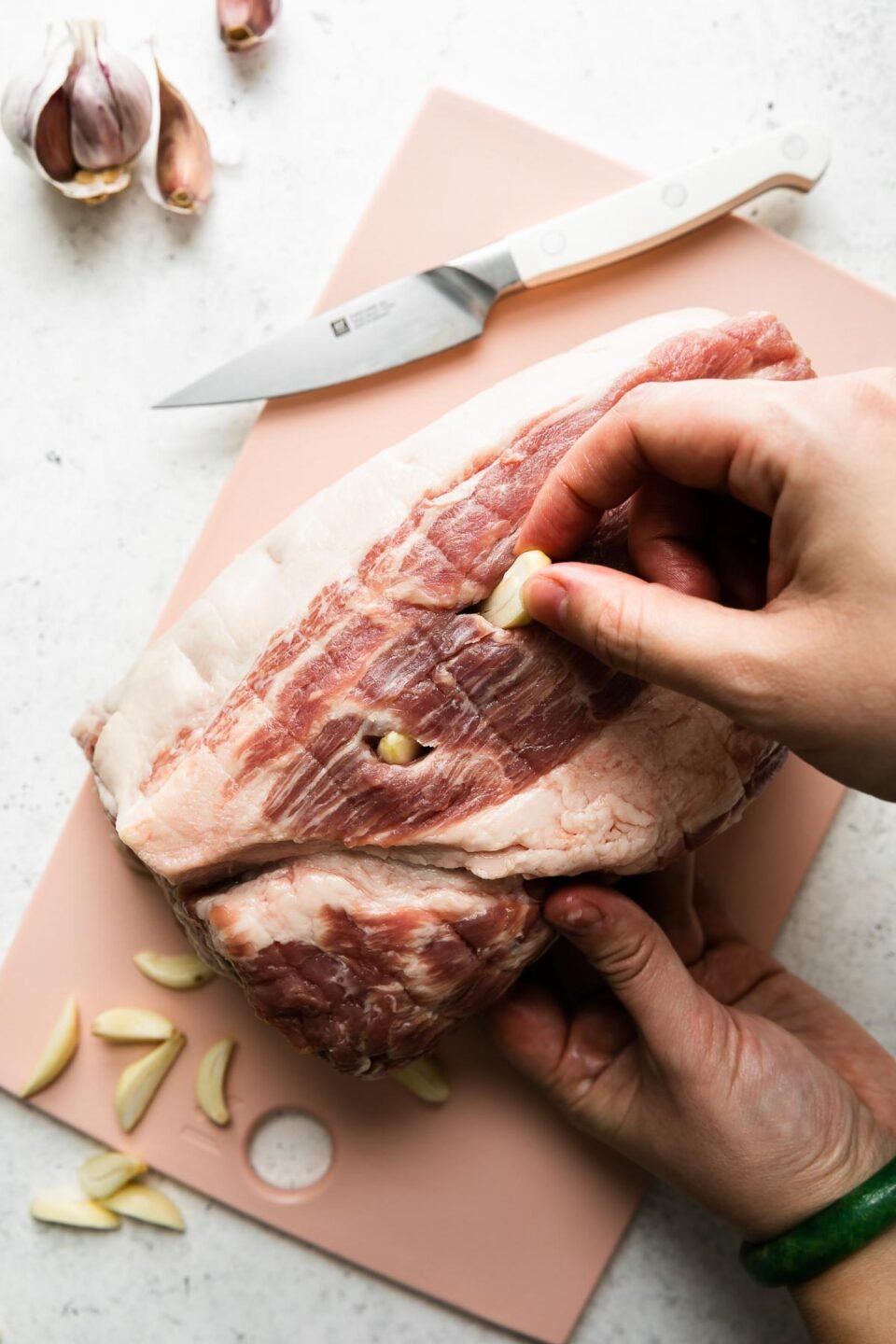 How to make braised carnitas, step 1: Stud & season the pork shoulder. A woman's hands work to insert sliced garlic cloves into slits made within a boneless pork shoulder. The pork shoulder sits atop a pink plastic cutting board that sits atop a creamy white textured surface. A pairing knife with a white handle rests on the cutting board along with a pile of loose peeled & sliced garlic cloves. Partial unpeeled garlic cloves rest alongside the cutting board.