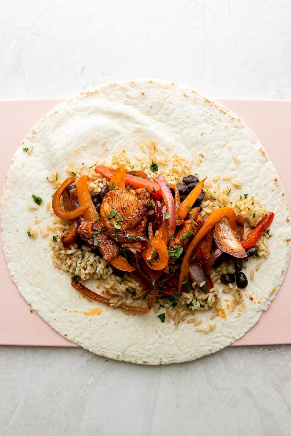 How to make chicken fajita burritos, step 5: Burrito assembly. A fajita burrito has been built atop a large flour tortilla. The tortilla sits flat on a pink plastic cutting board and the cutting board sits atop a creamy white textured surface.