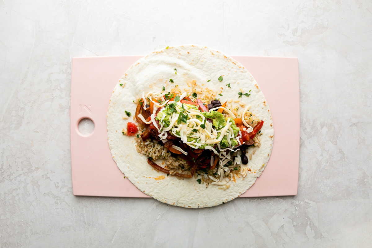 How to make chicken fajita burritos, step 5: Burrito assembly. A fajita burrito has been built atop a large flour tortilla and toppings have been added. The tortilla sits flat on a pink plastic cutting board and the cutting board sits atop a creamy white textured surface.