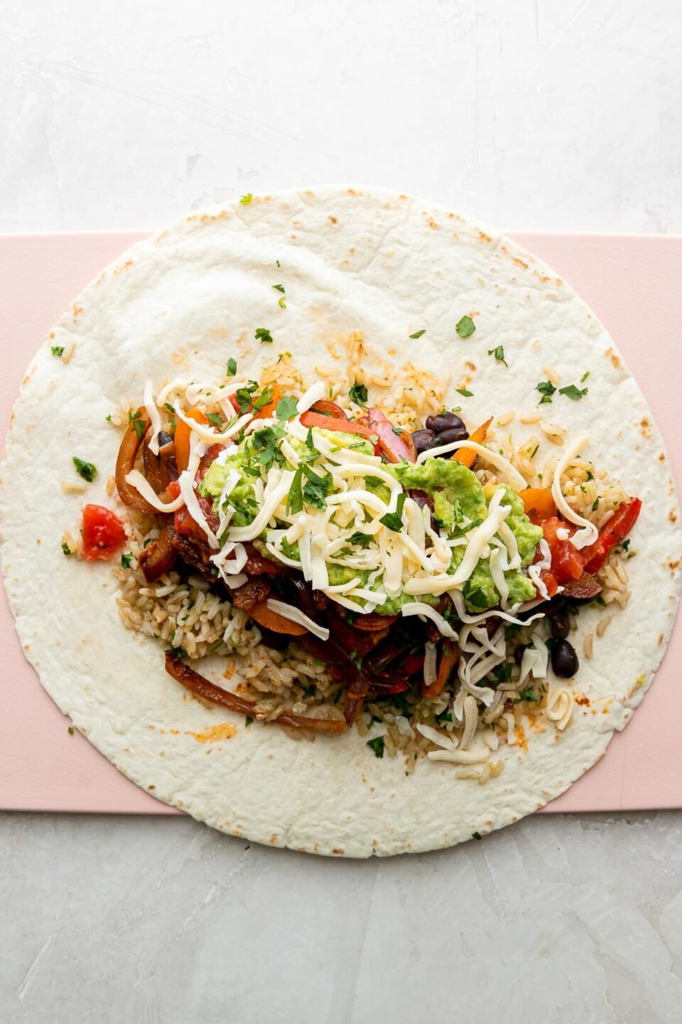How to make chicken fajita burritos, step 5: Burrito assembly. A fajita burrito has been built atop a large flour tortilla and toppings have been added. The tortilla sits flat on a pink plastic cutting board and the cutting board sits atop a creamy white textured surface.