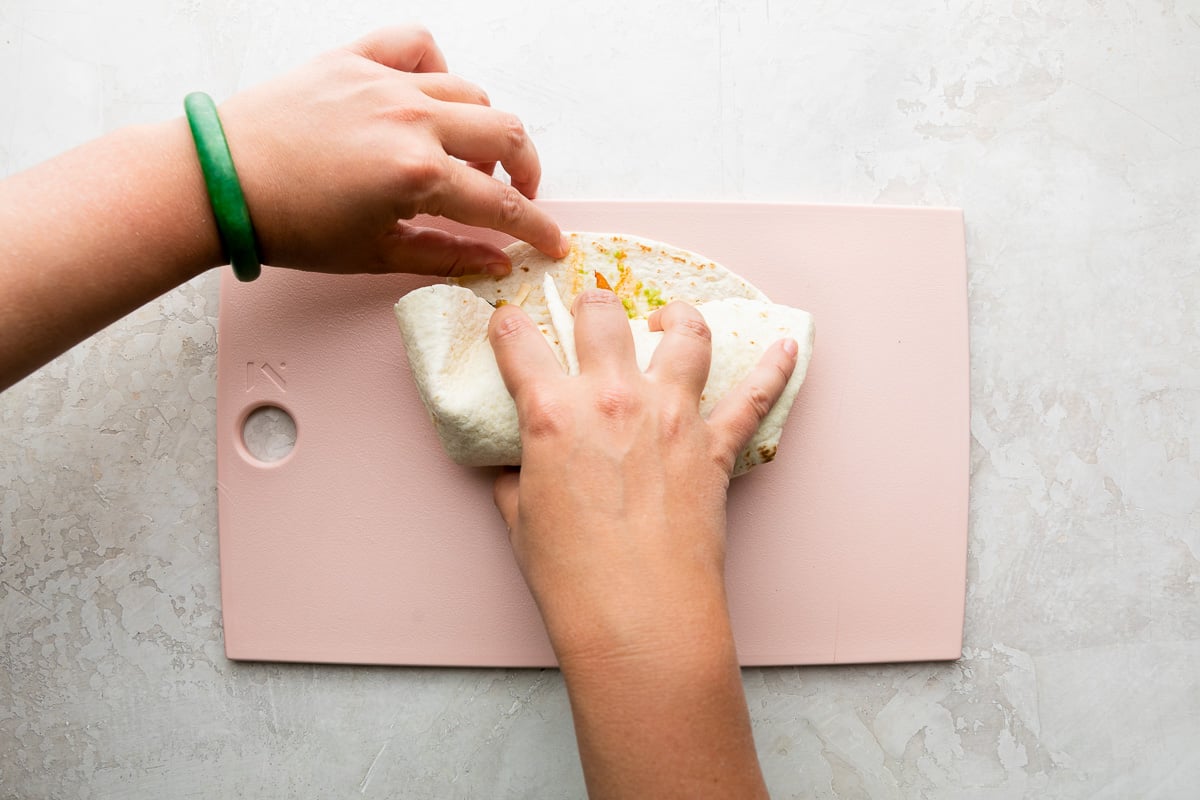 How to make chicken fajita burritos, step 5: Burrito assembly. A fajita burrito has been built atop a large flour tortilla with toppings added. The tortilla sits flat on a pink plastic cutting board and the cutting board sits atop a creamy white textured surface. A woman's hands work to roll the burrito.