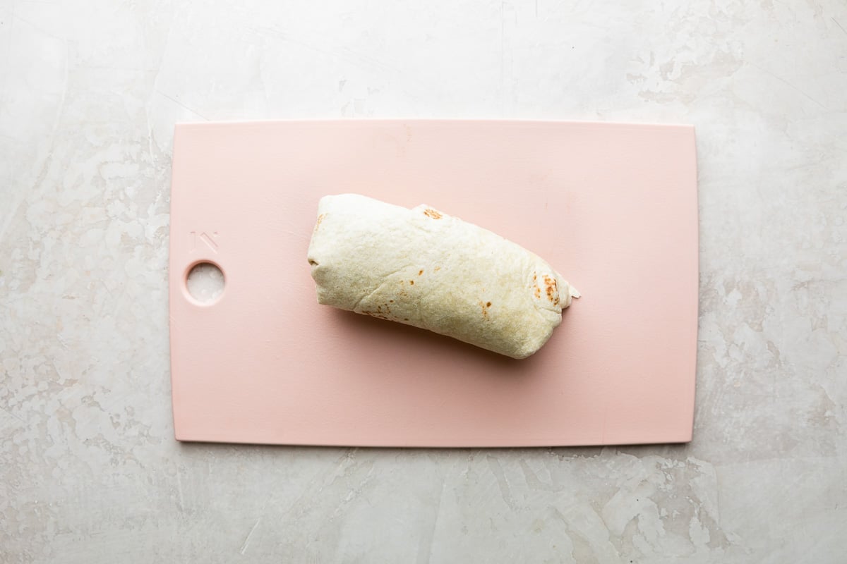 How to make chicken fajita burritos, step 5: Burrito assembly. A rolled chicken fajita burrito sits atop a pink plastic cutting board. The cutting board sits atop a creamy white textured surface.