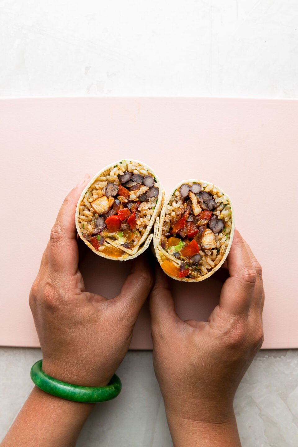 A woman's hands hold a finished chicken fajita burrito that has been cut in half. The burrito halves rest atop a pink plastic cutting board with the filling sit facing up to reveal what is inside. The cutting board sits atop a creamy white textured surface.