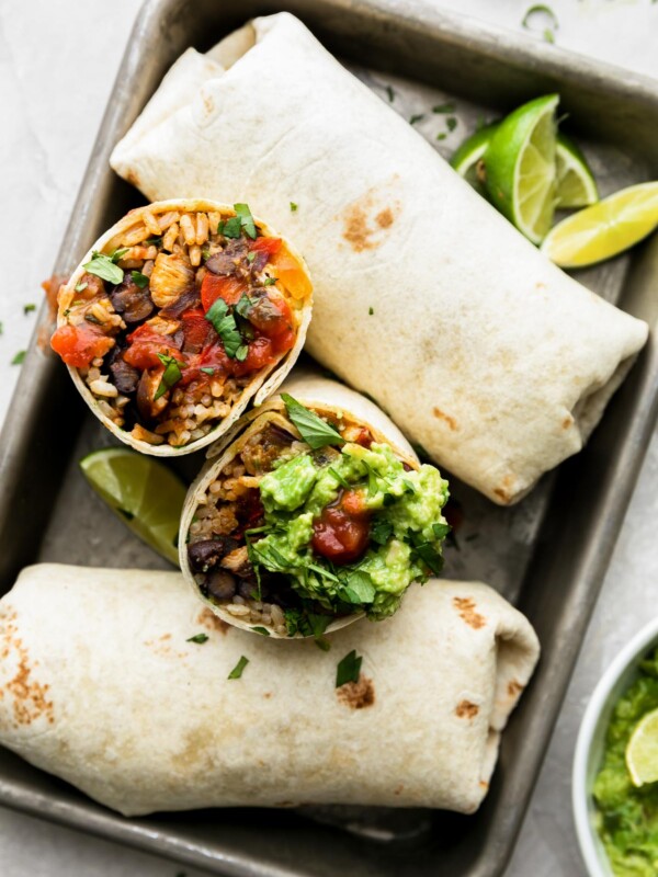 Finished chicken fajita burritos made with ingredients from ALDI sits atop small aluminum baking sheet. Two of the burritos are whole, while one of the burritos, at center, has been cut in half and rests with the filling side facing upwards. Limes wedges are tucked alongside the burritos and also surround the baking sheet. A small white ceramic bowl is filled with mashed avocado and all items sit atop a creamy white textured surface.